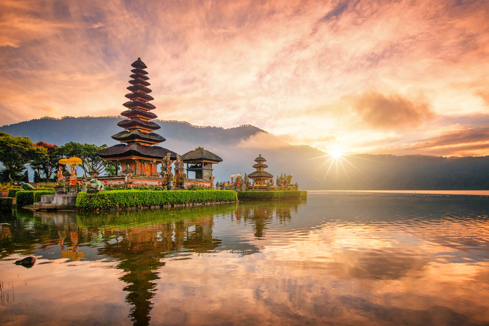 The same lake temple at sunrise, its tiered roofs dark against an orange sky, with the sun breaking over the ridge behind it.