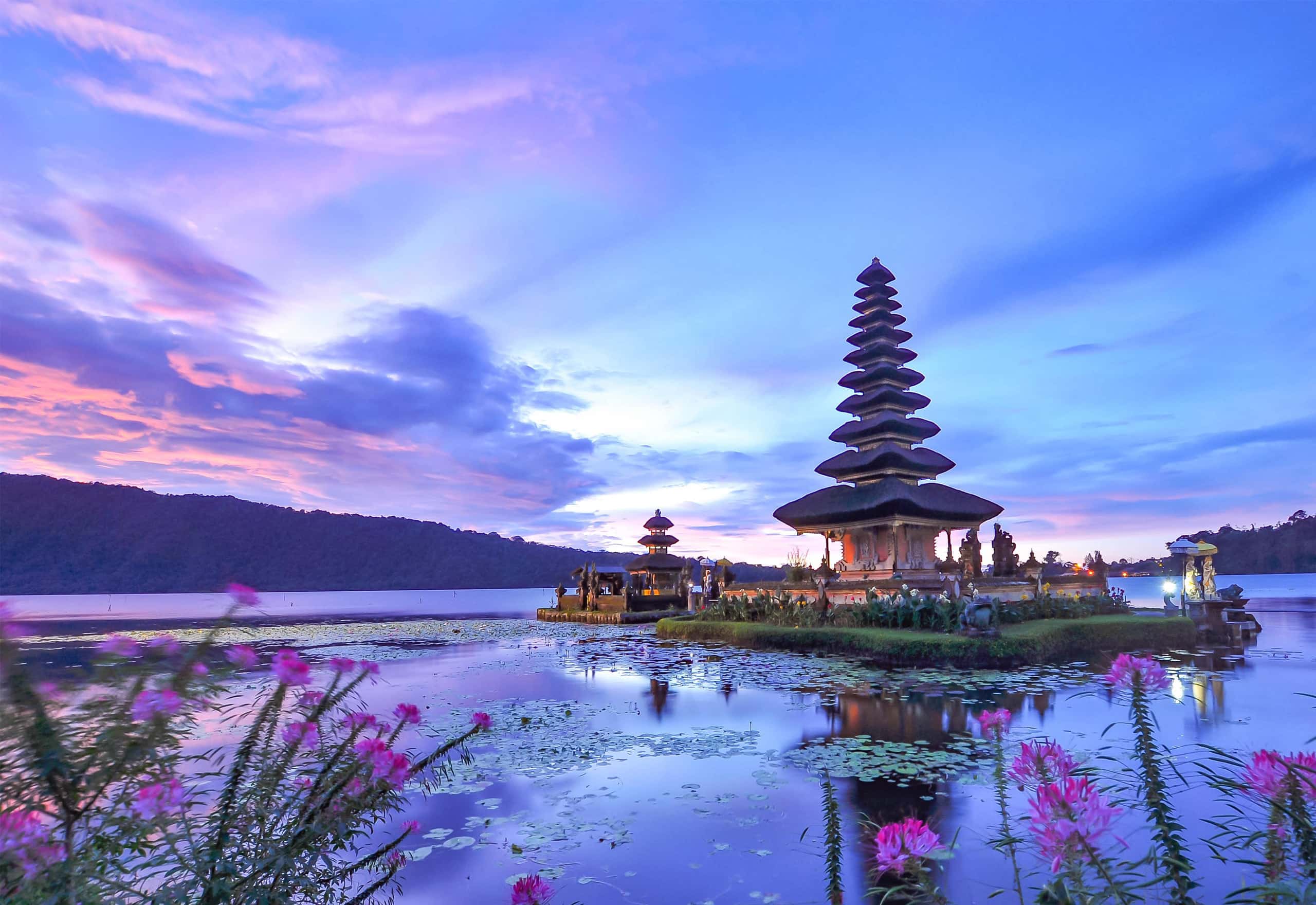 A tiered Balinese temple standing on the water of Lake Beratan at dusk, with pink and violet cloud reflected in the still lake.